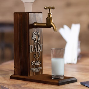 May include: A wooden Raki dispenser with a brass faucet and a glass of Raki. The dispenser has the word "RAKI" engraved on it, along with a fish emblem and the name "PETER MILLER". The dispenser is made of dark brown wood.