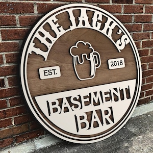 Puede incluir: Un letrero redondo de madera con el texto "The Jager's Basement Bar Est. 2018" en letras blancas. El letrero tiene un fondo de madera marrón y un borde blanco. Una silueta blanca de una jarra de cerveza está en el centro del letrero.