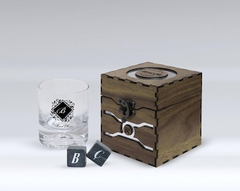 May include: A wooden box with a glass lid containing black whiskey stones. The box has a metal latch and a clear window showing the stones. A clear glass with a black and white logo is next to the box. Three black stones with white letters are in front of the box.