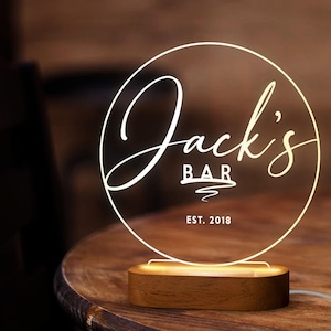 May include: A white illuminated sign with the text "Jack's Bar Est. 2018" in a circle, sitting on a wooden base.