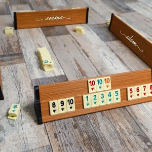 Puede incluir: Un juego de Rummikub de madera con fichas numeradas del 1 al 10. Las fichas están dispuestas en línea sobre un soporte de madera. El soporte está etiquetado con "adam" en un lado y "emma" en el otro. Las fichas son una mezcla de amarillo, azul y rojo.