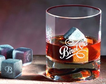 May include: A glass of whiskey with ice cubes and the words "Biroi Oil" engraved on the side of the glass.  There are several gray stone ice cubes on a wooden surface.