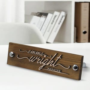 May include: A brown wooden desk nameplate with the text "emma wright MANAGER" in a cursive font. The nameplate has two silver screws on the top and one on the bottom.