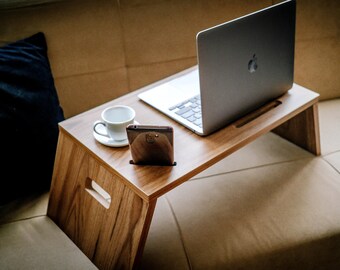 May include: A wooden laptop stand with a laptop, a smartphone, and a cup of coffee on it. The stand is on a light brown couch.