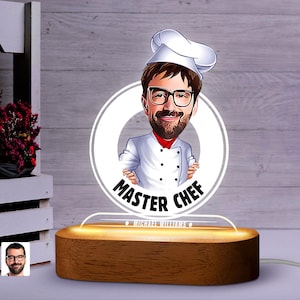 May include: A personalised night light featuring a cartoon illustration of a chef wearing a white chef's hat and a white chef's uniform. The illustration is on a white circle with the text "Master Chef" in black lettering. The night light is on a wooden base with the name "Michael Williams" in black lettering.