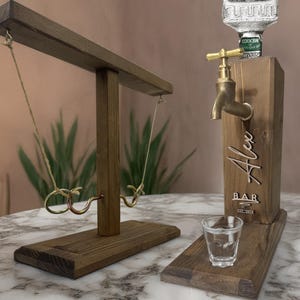 May include: A wooden bar game set with a ring toss game and a liquor dispenser. The dispenser has a brass faucet and the name "Alex" engraved on the front. A shot glass sits on the base.