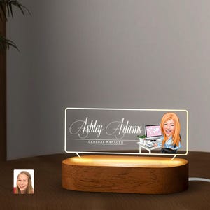 以下が含まれることがあります： 机の上に置かれた、女性がデスクで仕事をしている様子が描かれた、漫画風のイラストが施された、アクリル製のパーソナルデスクランプ。ランプは木製の台座に置かれており、"Ashley Adams, General Manager"という文字が書かれている。