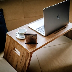 Puede incluir: Un soporte de madera para portátil con un portátil, un teléfono inteligente y una taza de café encima. El soporte está sobre un sofá de color marrón claro.