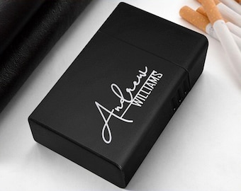May include: A black cigarette case with the name "Andrew Williams" in white script. The case is rectangular and has a matte finish. Cigarettes are visible in the background.