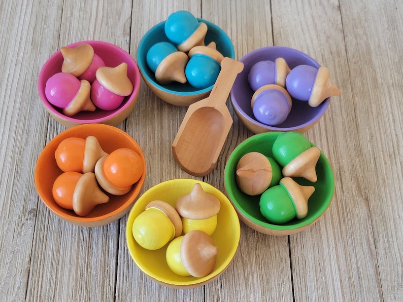 May include: Six wooden bowls filled with colourful acorn-shaped wooden toys. The bowls are arranged in a circle on a wooden surface. A wooden scoop is in the centre of the circle.
