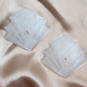 May include: Two clear, fan-shaped decorative pieces with a small hole in the center of each. The pieces are made of a translucent material and have a layered design. The background is a light, neutral color.