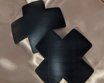 May include: Two black adhesive fabric pasties in the shape of a cross. The pasties are on a light brown fabric background.