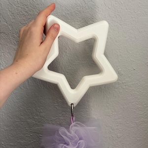 May include: A white star-shaped wall hook with a ring attached to a purple loofah. The hook is held by a hand. The star has a thick, layered design. The background is a textured gray wall.