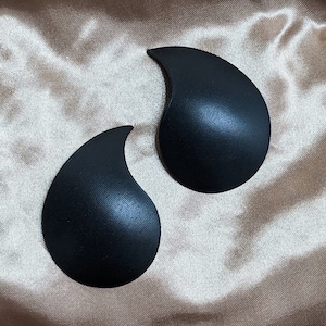May include: Two black, teardrop-shaped bra inserts. The inserts have a textured surface and a curved top. The background is a light brown, satin-like fabric.