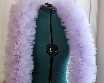 May include: A light purple tulle bolero jacket with ruffled trim, displayed on a green dress form. The bolero has long sleeves and a wide, ruffled collar. The tulle fabric is layered, creating a voluminous effect. The dress form has adjustable dials.
