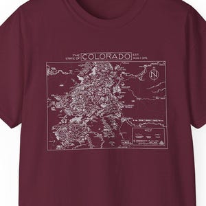 May include: A maroon t-shirt with a white outline of a map of Colorado. The map includes the text "THE STATE OF COLORADO" and a compass rose.