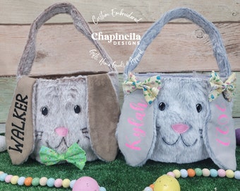 May include: Two gray bunny-shaped Easter baskets with brown ears. The basket on the left has a green bow and the name "WALKER" embroidered on the ear. The basket on the right has a yellow bow and the name "KYLAH" embroidered on the ear. The baskets are sitting on a green surface with colorful beads and Easter eggs.