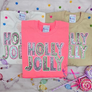 May include: Three pink, white, and beige t-shirts with the words "HOLLY JOLLY" and "HOLLY" in silver sequins on the front. The shirts are laid out on a surface with candy and a wand.