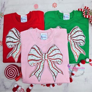May include: Three Christmas-themed t-shirts with a sparkly red and green bow design. The shirts are red, green, and pink.