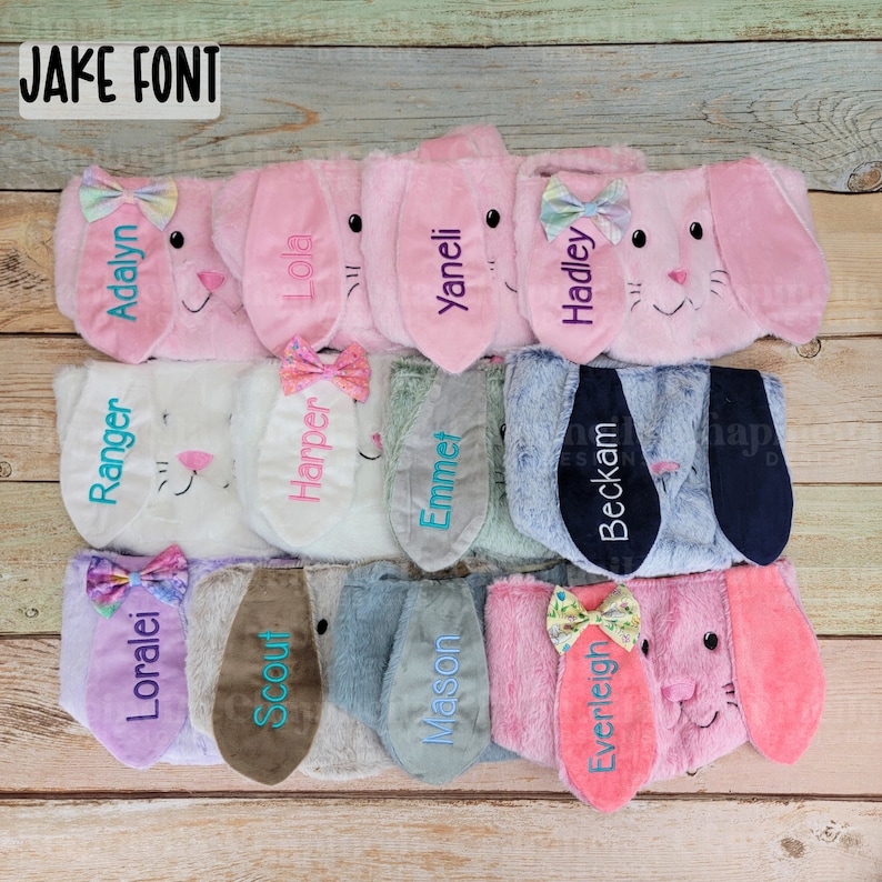 Puede incluir: Una colecci&oacute;n de diademas de orejas de conejo de felpa en varios colores, incluyendo rosa, blanco y azul. Cada diadema presenta nombres bordados y un lazo decorativo. El texto "JAKE FONT" est&aacute; en la esquina superior izquierda.