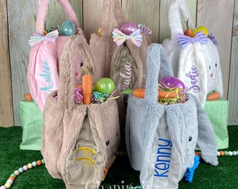 Peut inclure: Quatre paniers de Pâques en forme de lapin en peluche avec des noms personnalisés, dont Aubrey, Julia, Sadie et Kenny. Chaque panier est rempli d'herbe de Pâques colorée, d'œufs en plastique et d'une carotte. Les paniers sont disposés sur une surface herbeuse verte avec un fond en bois.