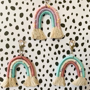 May include: Three handmade rainbow keychains. Each keychain features a macrame rainbow design in pastel colors: pink, blue, and purple. The rainbows have a fringe at the bottom and are attached to a silver clasp.