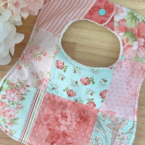 May include: A patchwork baby bib with a variety of floral and striped patterns in shades of pink, peach, and mint green. The bib has a rounded neck opening and a snap closure. The bib is made of soft fabric.