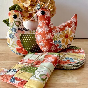 May include: Two stuffed fabric chickens with floral and polka dot patterns in orange, green, yellow, and white. One chicken is primarily orange with white flowers, the other is white with colorful floral prints. Includes a stack of quilted fabric coasters.