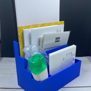 May include: A blue plastic organizer with multiple compartments for storing craft supplies. It contains paper, envelopes, and a white dispenser with a green lid.