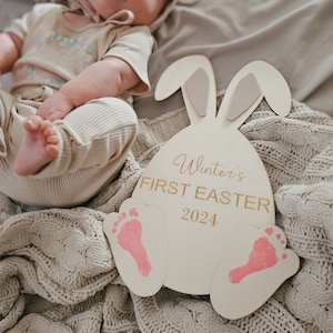 May include: A wooden bunny-shaped sign with pink footprints and the text "Winter's First Easter 2024". A baby is lying on a knitted blanket in the background.