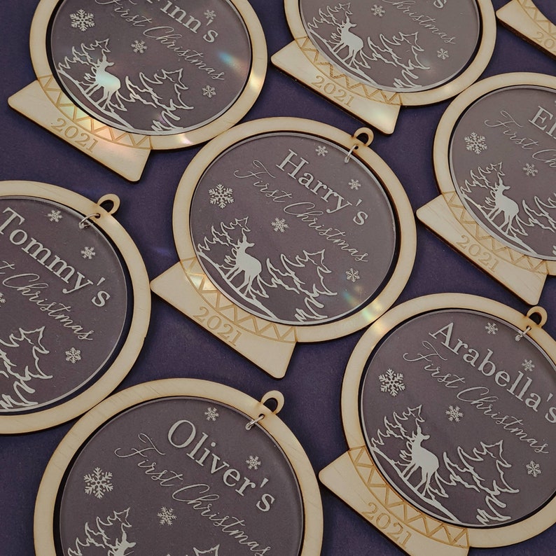 May include: A set of six wooden ornaments with clear acrylic inserts. Each ornament features a reindeer and trees in a snowy forest scene. The ornaments are personalized with the names "Harry's", "Tommy's", "Oliver's", "Arabella's", "Inn's", and "El's" and the year "2021".