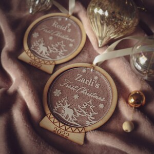May include: Two personalized wooden Christmas ornaments with a snow globe design. The ornaments are decorated with a reindeer, trees, and snowflakes. The text on the ornaments reads "Reid's First Christmas 2022" and "Zarli's First Christmas 2022".