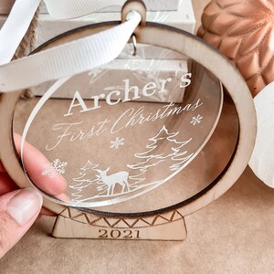 May include: A clear round ornament with a wooden frame and stand. The ornament is engraved with the text "Archer's First Christmas" and the year "2021". The ornament features a deer and trees in a winter scene.
