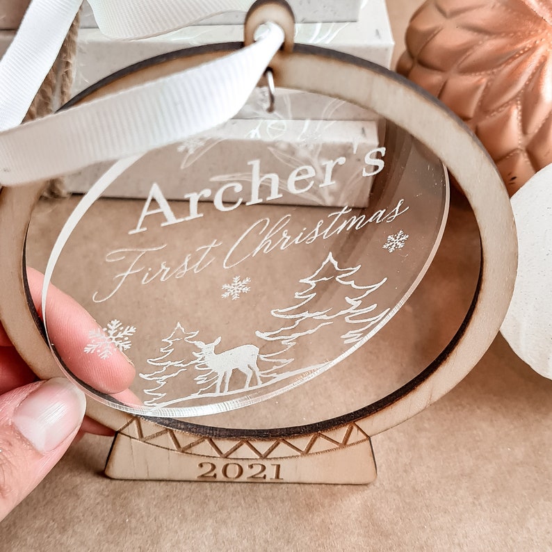May include: A clear round ornament with a wooden frame and stand. The ornament is engraved with the text "Archer's First Christmas" and the year "2021". The ornament features a deer and trees in a winter scene.