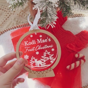 May include: A round, wooden Christmas ornament with a clear acrylic insert. The ornament features a white silhouette of a winter scene with trees and a deer. The text "KODI-MAE'S FIRST CHRISTMAS" is written in white on a red background. The year "2023" is written on the bottom of the ornament.