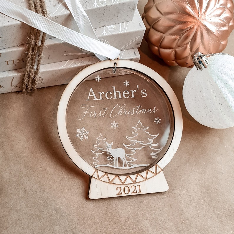 May include: A wooden Christmas ornament with a clear acrylic insert featuring a deer and trees. The ornament is engraved with the text "Archer's First Christmas 2021".