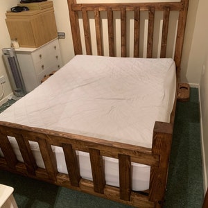 May include: A dark stained wooden bed frame with a slatted headboard and footboard. The bed has a white mattress and bedding. The bed frame is made of thick, solid wood. The bed is a double, king or super king size, depending on the dimensions.