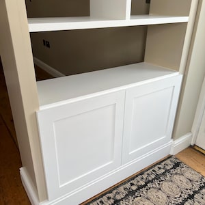 May include: White built-in bookshelf with two closed cabinets at the bottom. The shelves are empty and the cabinets have a simple design with a raised panel.