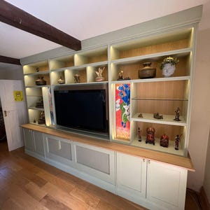 May include: A bespoke entertainment unit in a pale green hue, featuring a large television, open shelving displaying ornaments, and lower cabinets. The shelves are lit, and the unit is set against a light wall and wooden flooring.