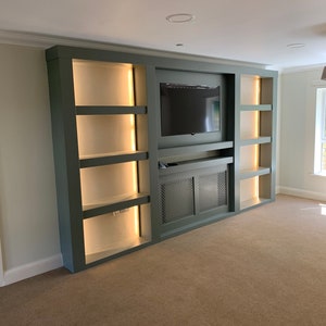 May include: A built-in entertainment unit with a dark green finish and a flat screen TV. The unit has shelves with warm white LED lights and a lower cabinet with a metal grille.