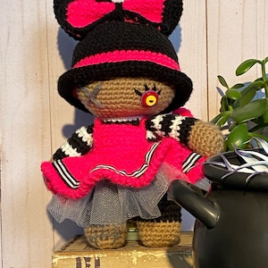 May include: A handmade crochet doll with a black hat and bow, accented with bright pink. The doll has a pink dress, a gray tulle skirt, and a single button eye. The doll is standing on a box with the word "FORTUNE" on it.
