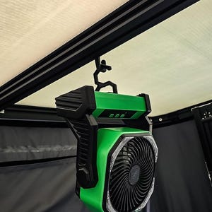 May include: A green and black portable fan is suspended from a black metal frame. The fan has a circular grill with a spiral design and a green body with black accents. The fan is designed for outdoor use.