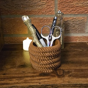 May include: A brown rope-textured desk organizer holds a pen, scissors, and a decorative wand with a crystal top. A black marker and a small glass bottle are also visible. The organizer sits on a wooden surface.