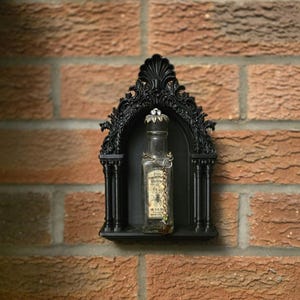 Peut inclure: Une étagère murale noire de style gothique avec un sommet en arc, contenant une bouteille en verre vintage. La bouteille a une étiquette décorative et un bouchon orné de bijoux. L'étagère est fixée sur un mur de briques rouges.