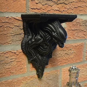 Può includere: Mensola nera con una scultura dettagliata di una creatura aliena. La mensola è montata su un muro di mattoni rossi. Il design alieno presenta una testa prominente e dettagli strutturati.