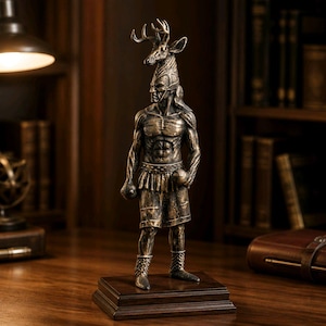 May include: Bronze statue of a muscular figure with a deer head headdress, holding two round objects. The statue stands on a dark wooden base. The figure is wearing shorts and boots. The statue is in a room with a desk and bookshelves.
