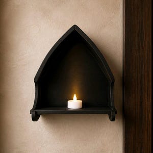 Pode incluir: Uma prateleira de madeira preta em forma de arco, montada numa parede bege, com uma pequena vela branca a arder no interior. A prateleira tem um design decorativo de inspiração gótica. O brilho quente da vela contrasta com a prateleira escura. Decoração de casa.