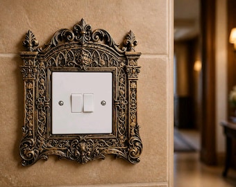 Puede incluir: Cubierta de interruptor de luz de estilo antiguo y ornamentado con un marco decorativo de color bronce. El marco rodea una placa de interruptor blanca con dos interruptores basculantes. El interruptor está montado en una pared de azulejos beige.
