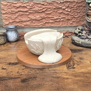 Op de afbeelding: Een witte keramische kom met een tarwe-aarontwerp, overvol met een witte, getextureerde substantie, staat op een ronde, bruine houten basis. De kom is aan de bovenkant open en de substantie stroomt eruit.