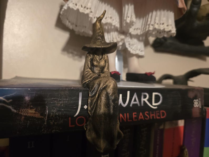 Puede incluir: Una figura de color bronce de un personaje sentado que lleva un sombrero puntiagudo y una t&uacute;nica larga y fluida. La figura est&aacute; colocada en el borde de un libro negro con el texto J. WARD LOVER UNLEASHED impreso en blanco y rojo en el lomo.
